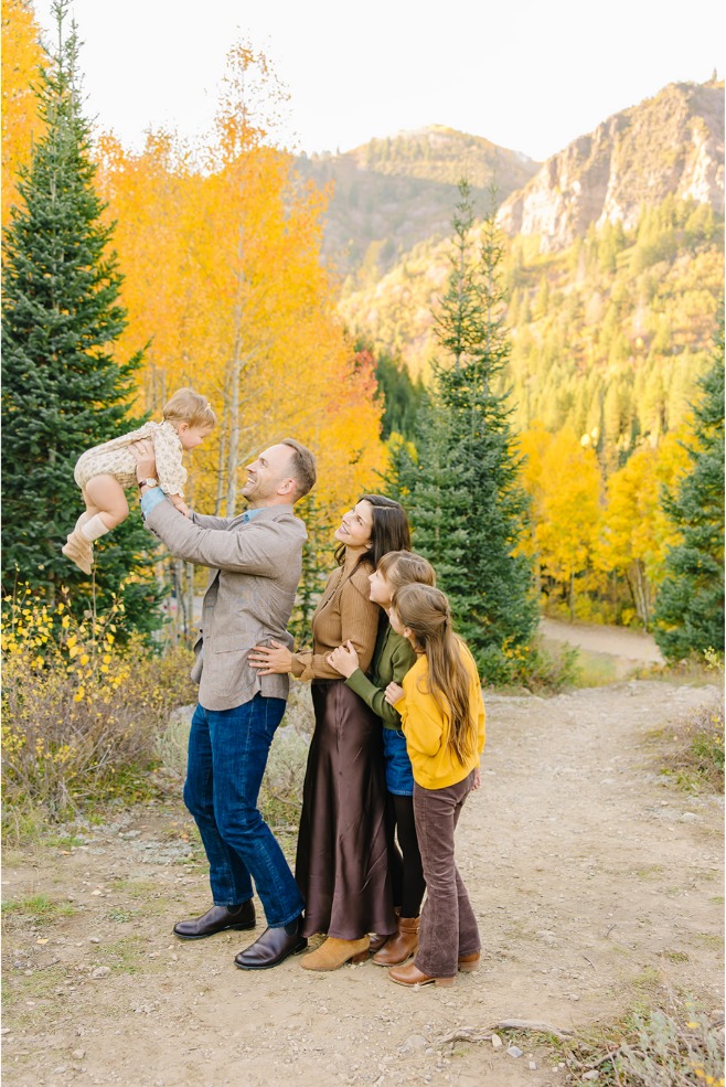 Dad lifting his baby girl into the air while mom and two daughters watch during fall family photos with baby in Utah