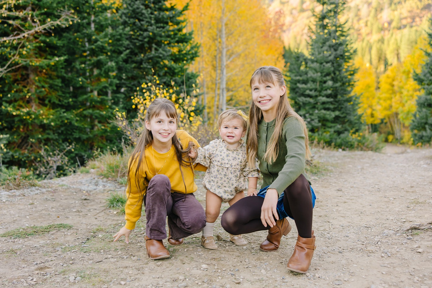 Two sisters crouched beside their baby sister and smiling among golden aspens and evergreens