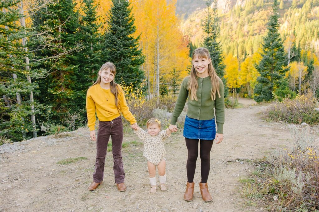 Two big sisters holding their baby sister's hands as she walks down a trail lined with fall foliage