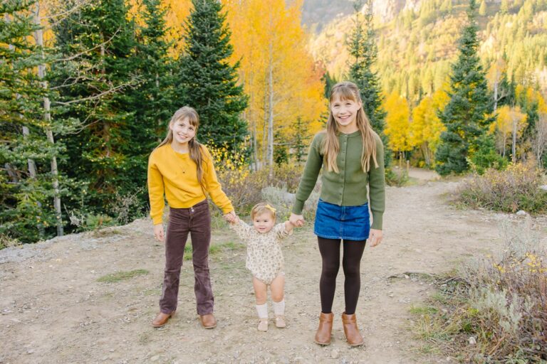 Two big sisters holding their baby sister's hands as she walks down a trail lined with fall foliage