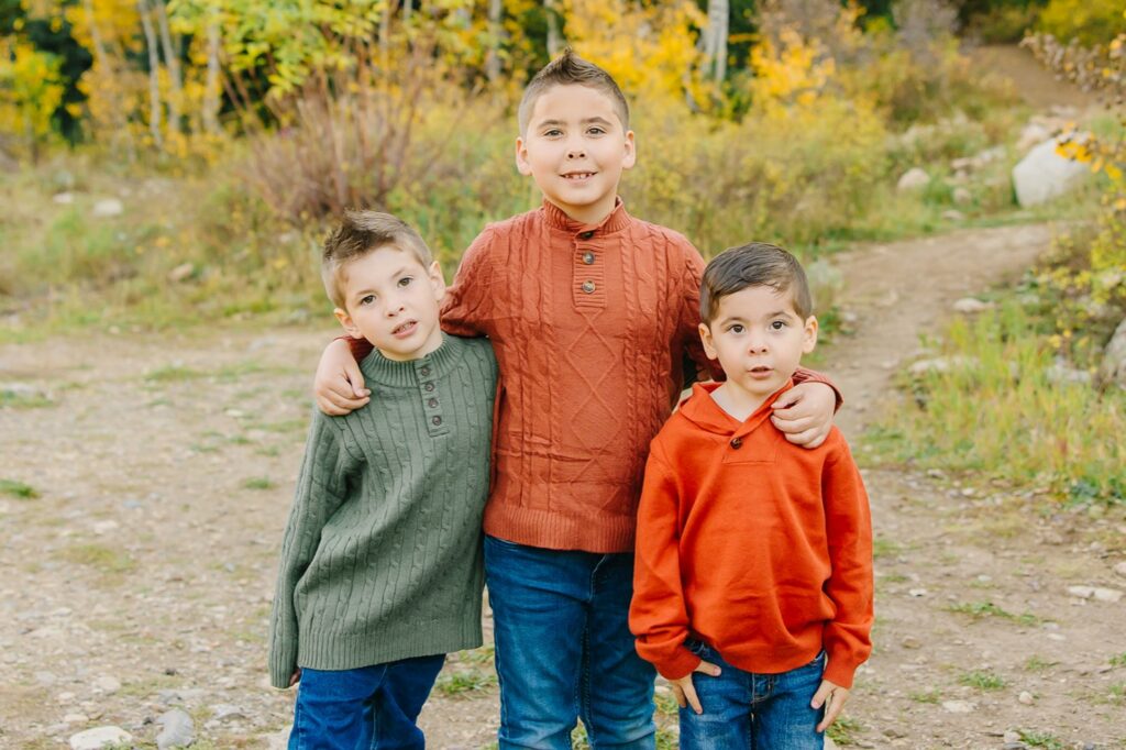 Three brothers in orange and sage green sweaters standing arm in arm on a dirt path during family photos with boys in Utah